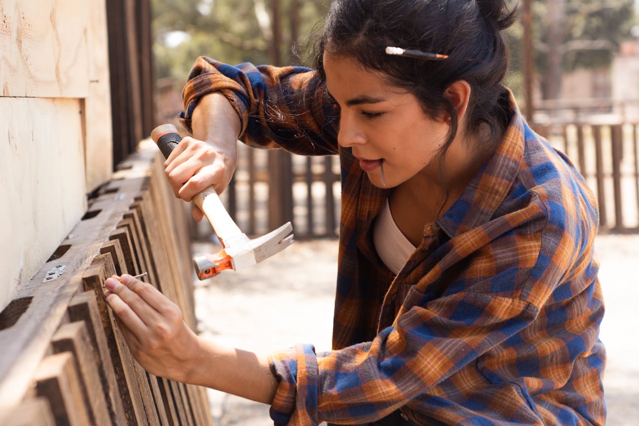 Young woman carpenter uses hammer to fix wooden fence outdoors.